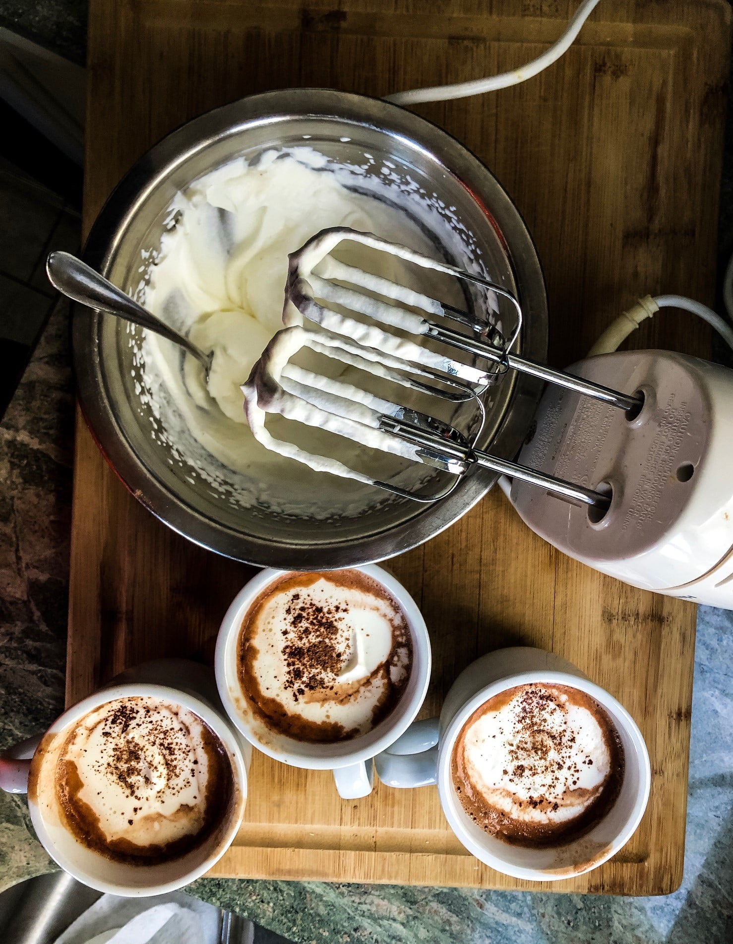 3 mugs of hot cocoa with a bowl of homemade whipped cream and hand mixer