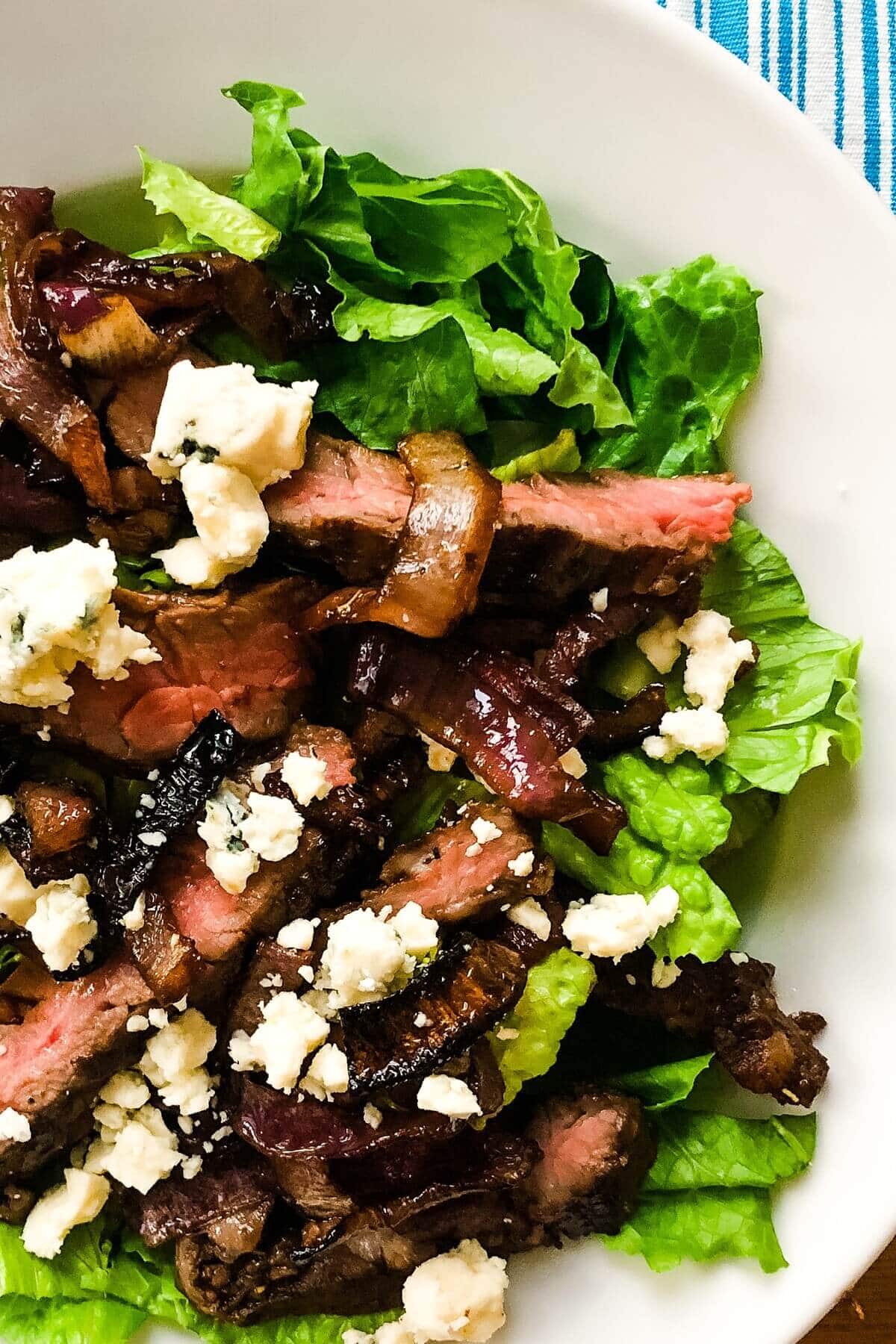 A bowl with lettuce, sliced steak, blue cheese and roasted onions.