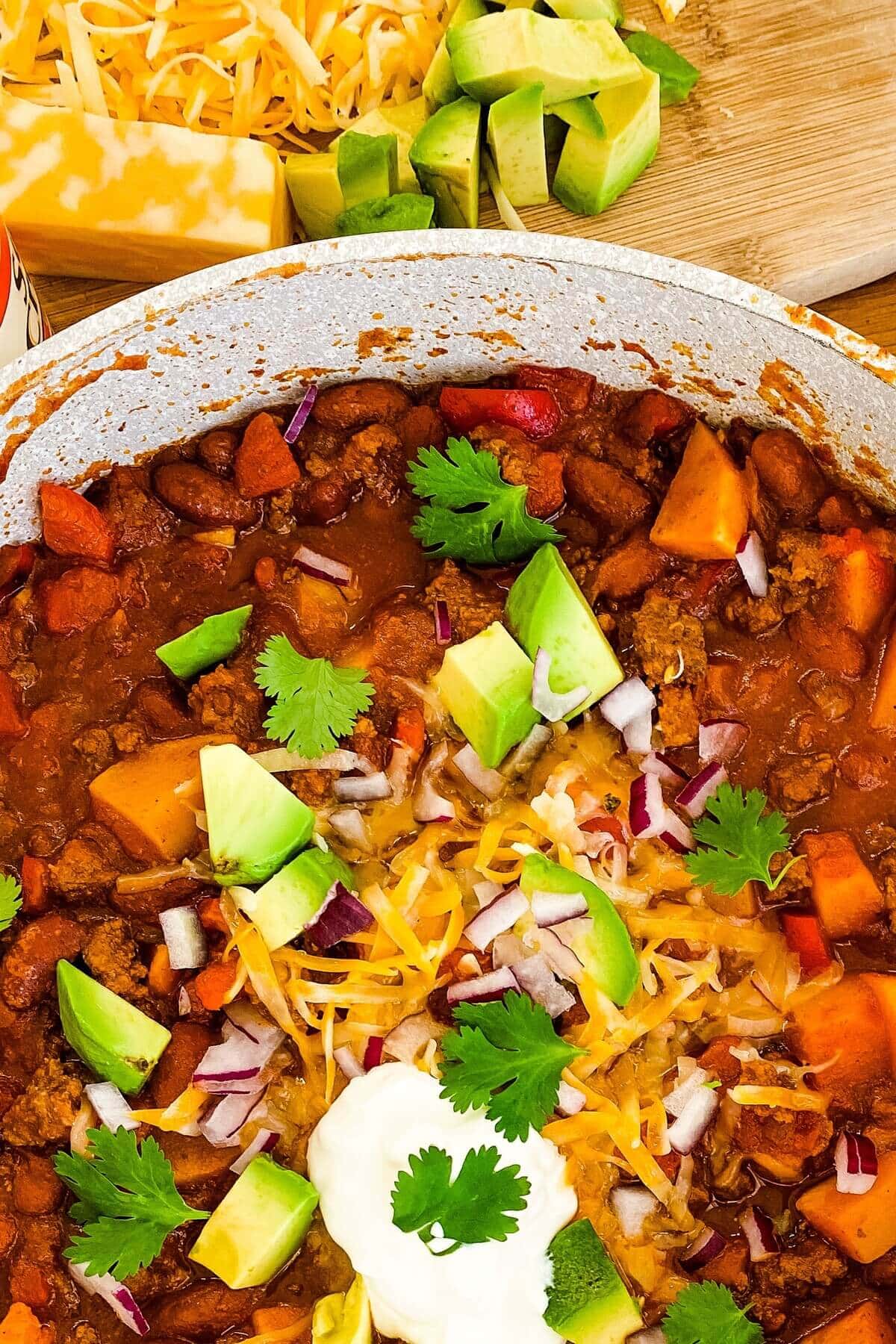 Chili with butternut squash in a pot topped with cheese, avocado and cilantro.