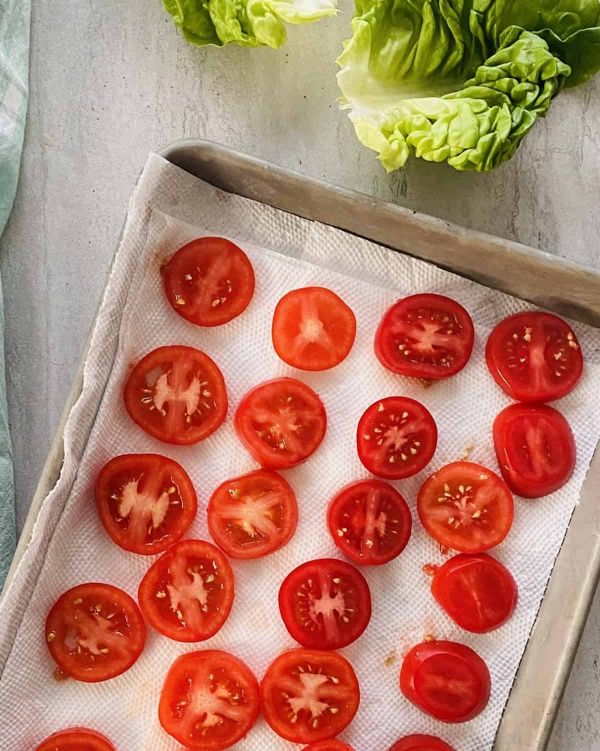 Slices of tomatoes draining on a paper towel lined baking sheet.