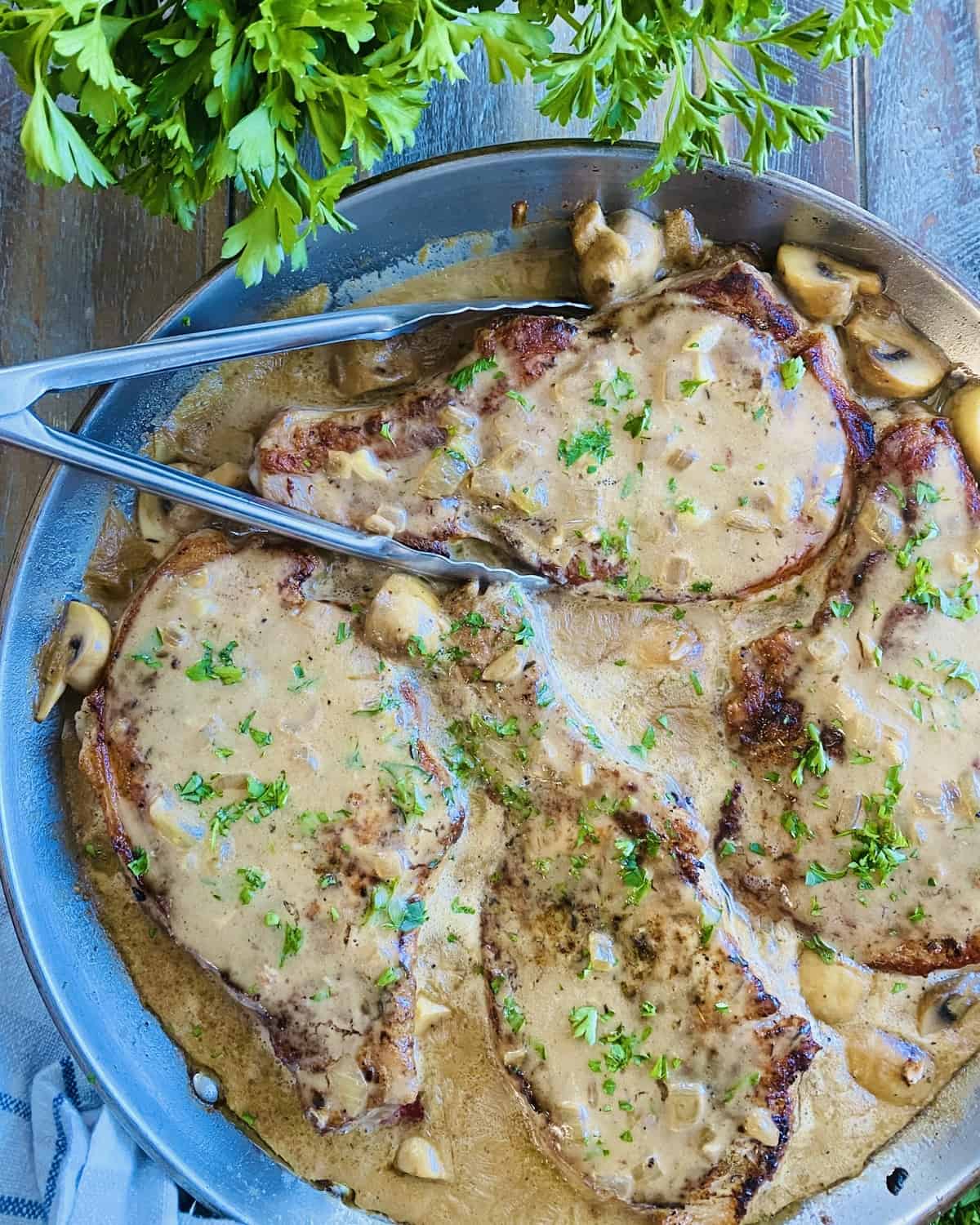 A pan of smothered pork chops with mushroom gravy and a pair of tongs.