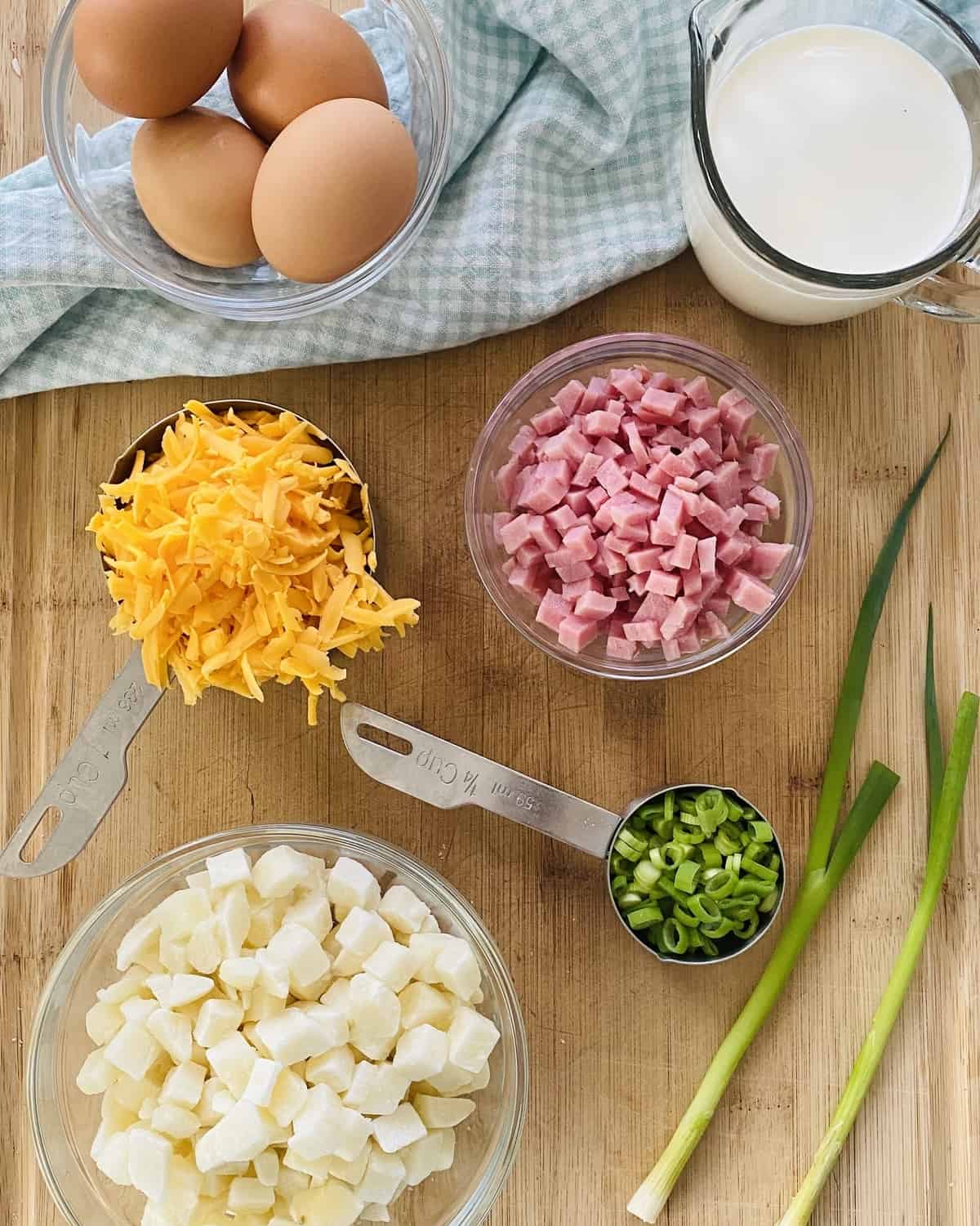 Eggs, ham, hash browns, heavy cream, cheddar cheese, scallions on a cutting board.