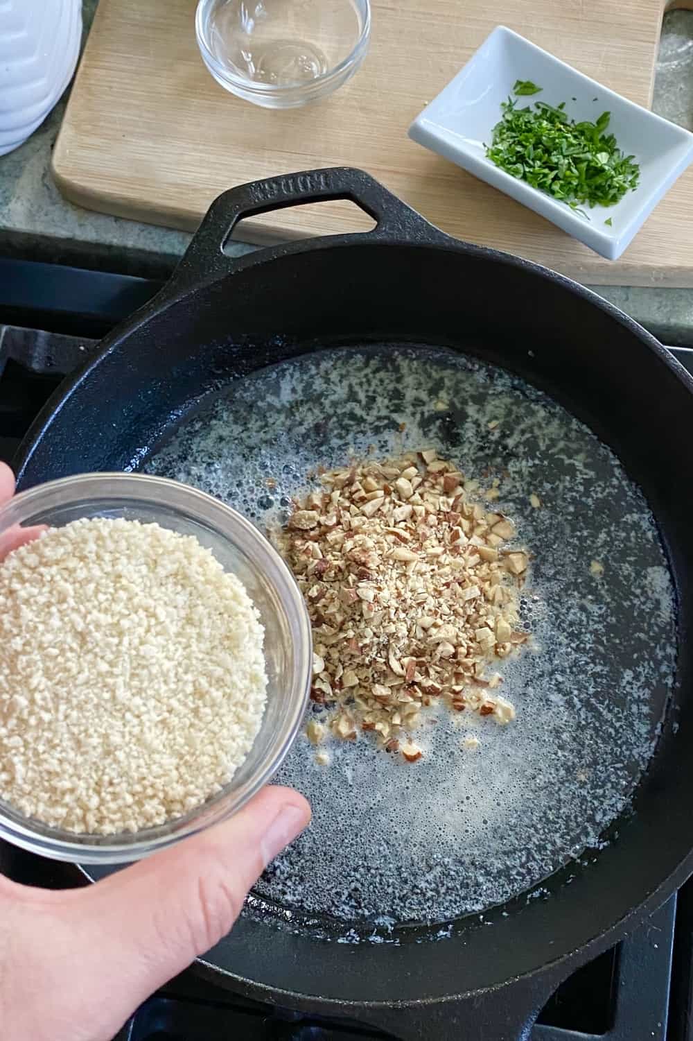 Adding panko to chopped pecans in a cast iron skillet.