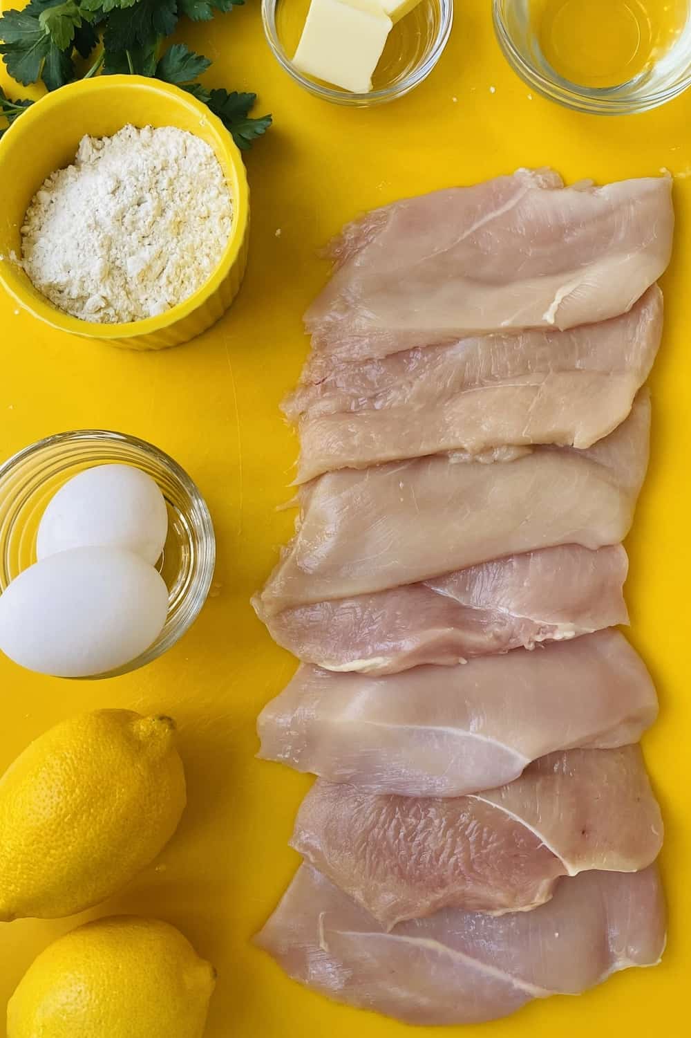 Chicken breasts, lemon, flour, white wine, and eggs on a yellow counter.