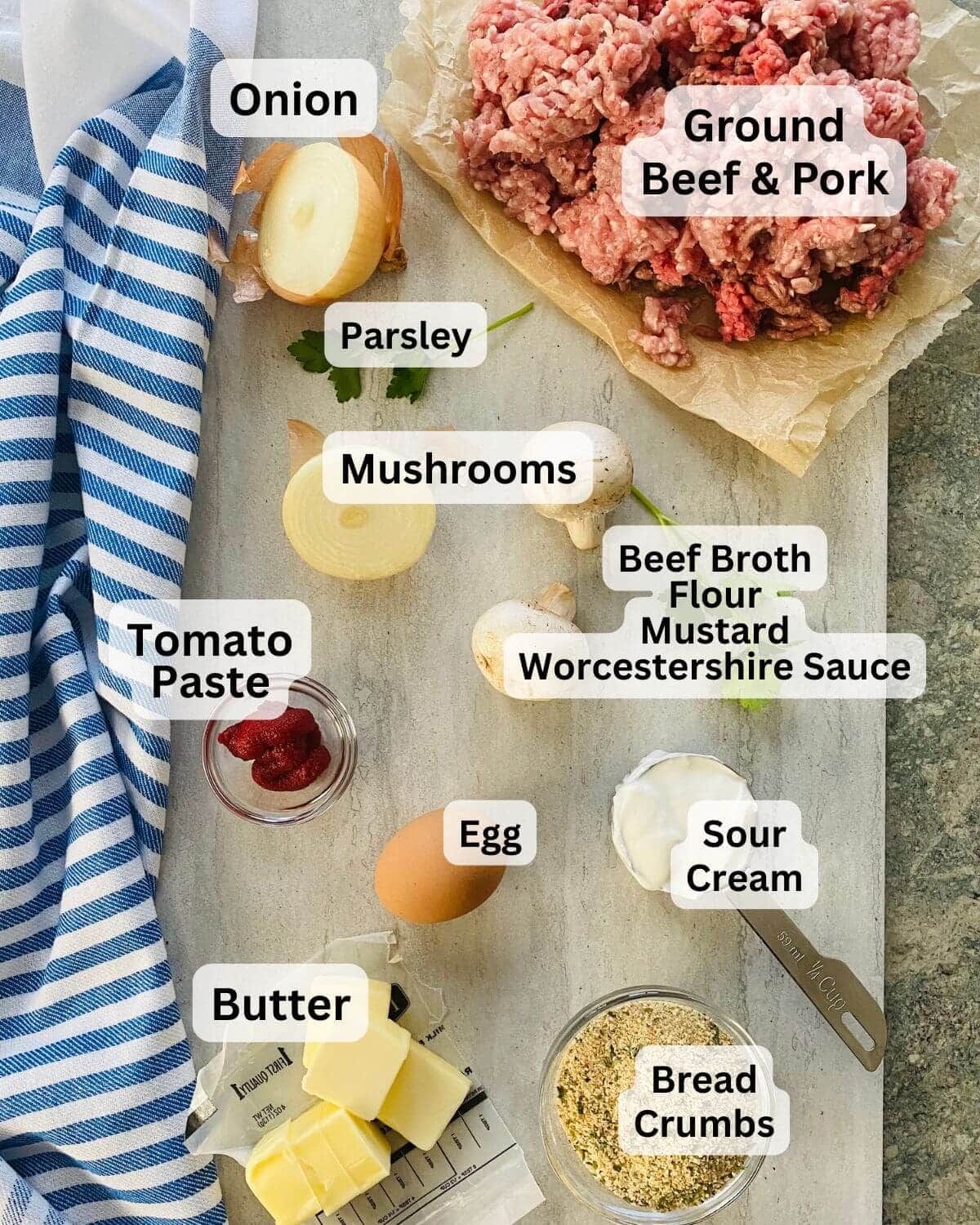 Ground beef and pork, onion, mushrooms, sour cream, parsley, egg, bread crumbs, and butter on a cutting board.