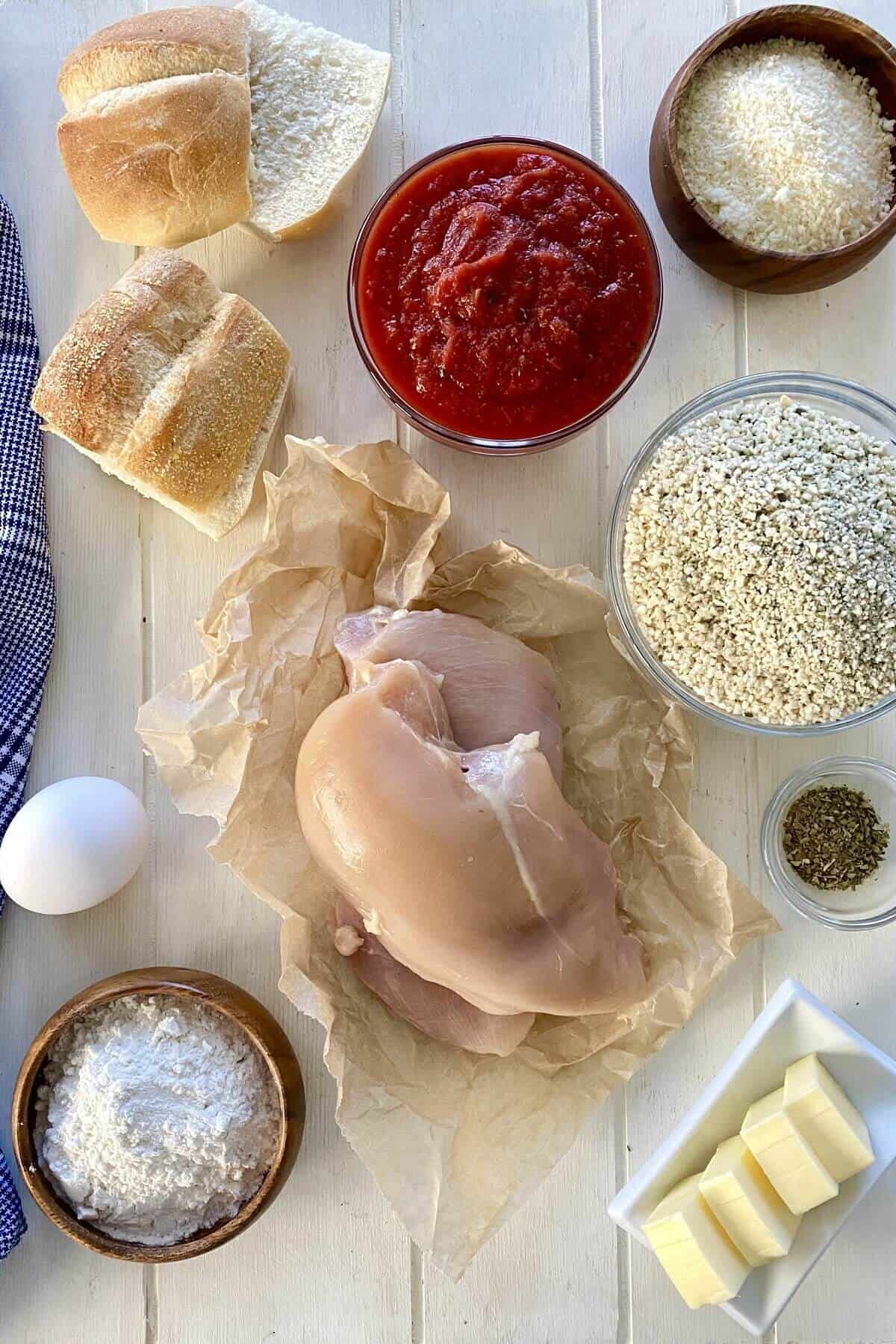 Chicken breast, marinara sauce, panko, egg, flour, rolls on a board.