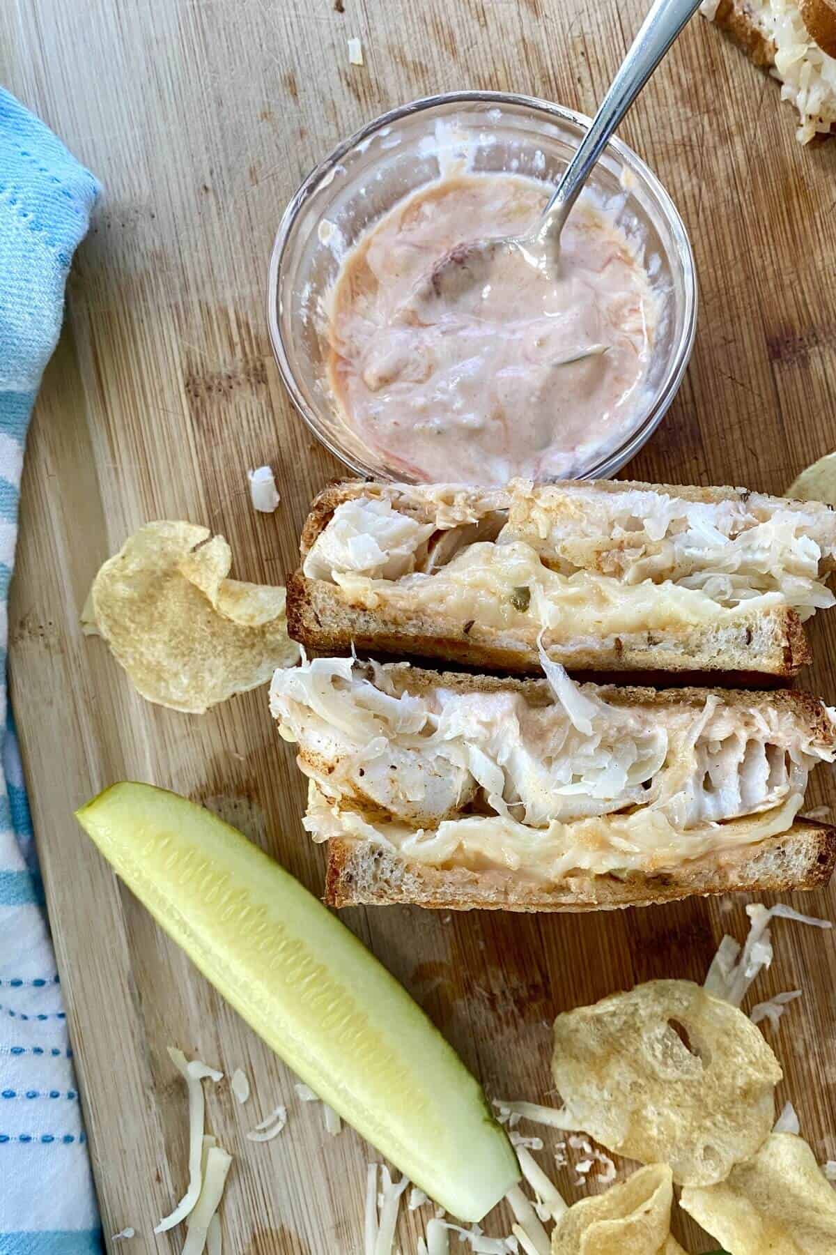 A cod fish reuben sandwich with a bowl of dressing.