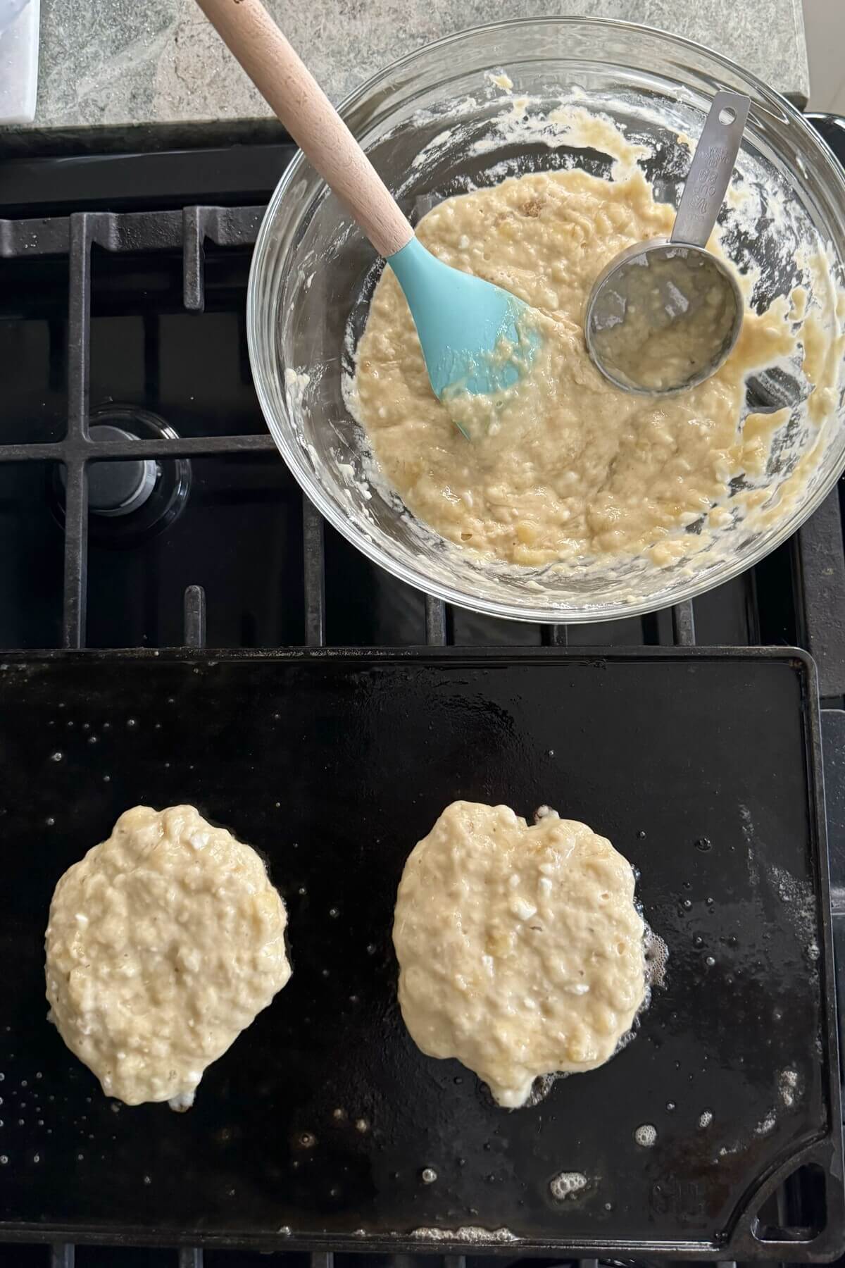 Pancake batter and 2 pancakes on a griddle.
