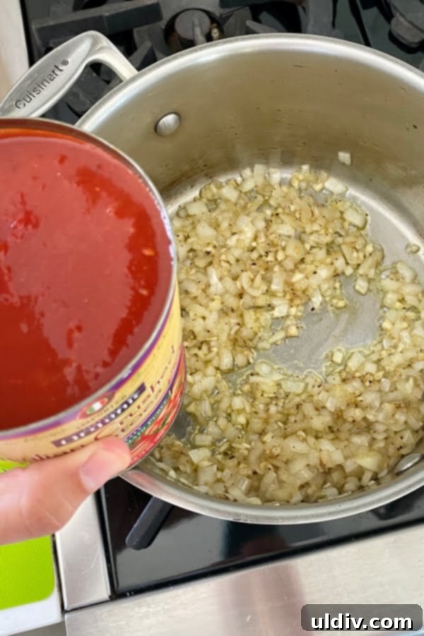 Signature Fresh Basil Marinara 3 Close-up of diced onions sautéing in olive oil in a saucepan, with a can of crushed tomatoes visible in the background, highlighting key marinara ingredients.