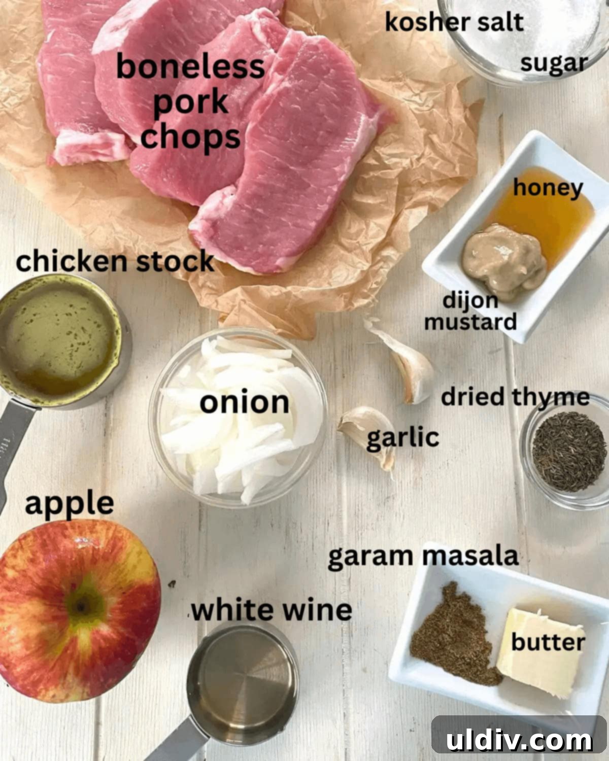 A flat lay of fresh ingredients including boneless pork chops, crisp apples, yellow onion, a block of butter, salt, mustard, and chicken stock, ready for preparation.