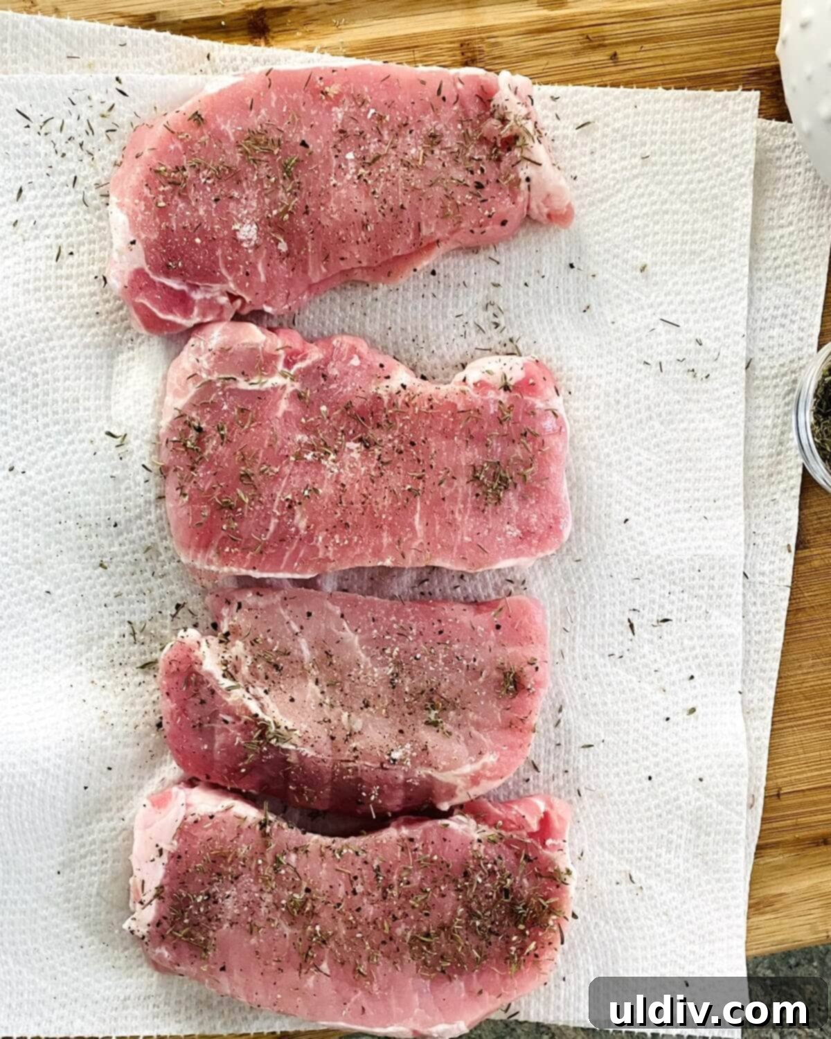 Close-up of raw pork chops being seasoned with dried thyme, kosher salt, and freshly ground black pepper on a cutting board.