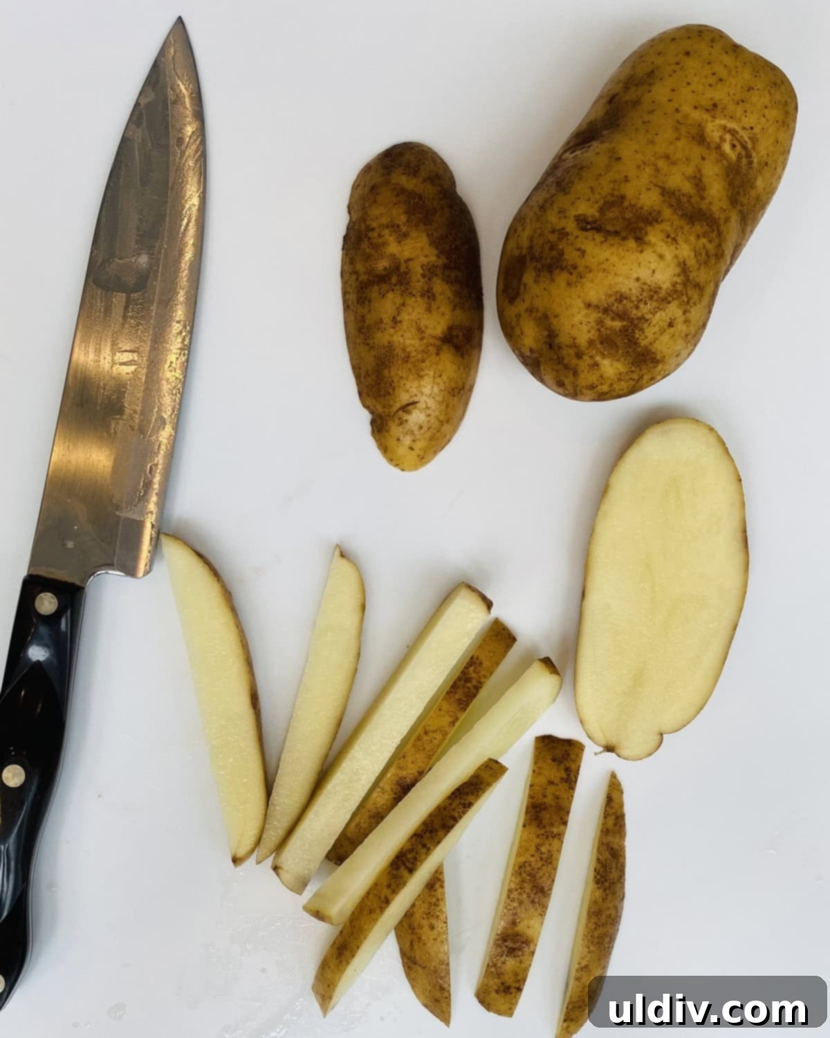 Homemade Crispy Oven Fries 4 A russet potato being expertly sliced into fry shapes on a cutting board with a chef's knife.