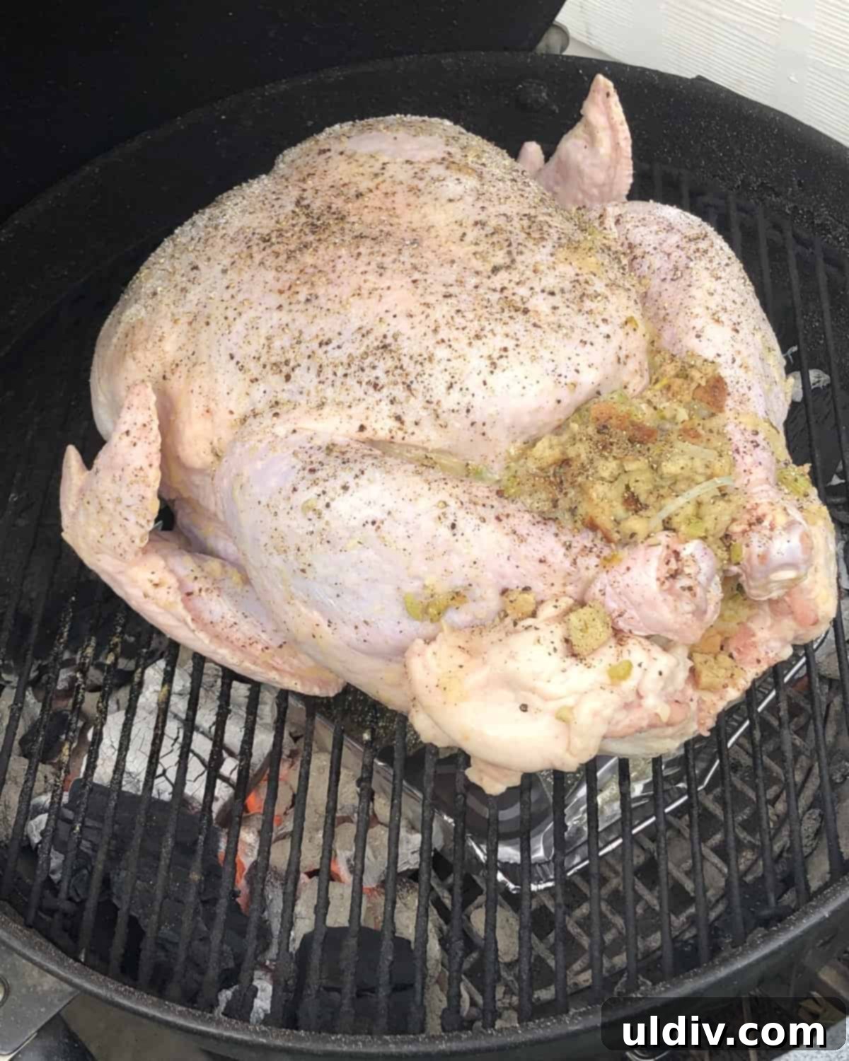 A whole turkey, glistening and golden brown, cooking over a drip pan on a hot charcoal grill, surrounded by briquettes.