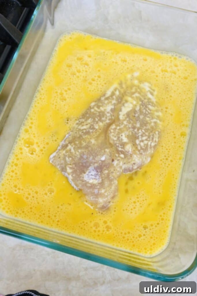 A hand dipping a floured chicken cutlet into an egg wash, preparing it for cooking.