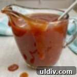 Simply Delicious Homemade BBQ Sauce 11 A clear glass pitcher filled with a rich, dark simple homemade BBQ sauce, garnished with a sprig of fresh herbs, ready for serving.