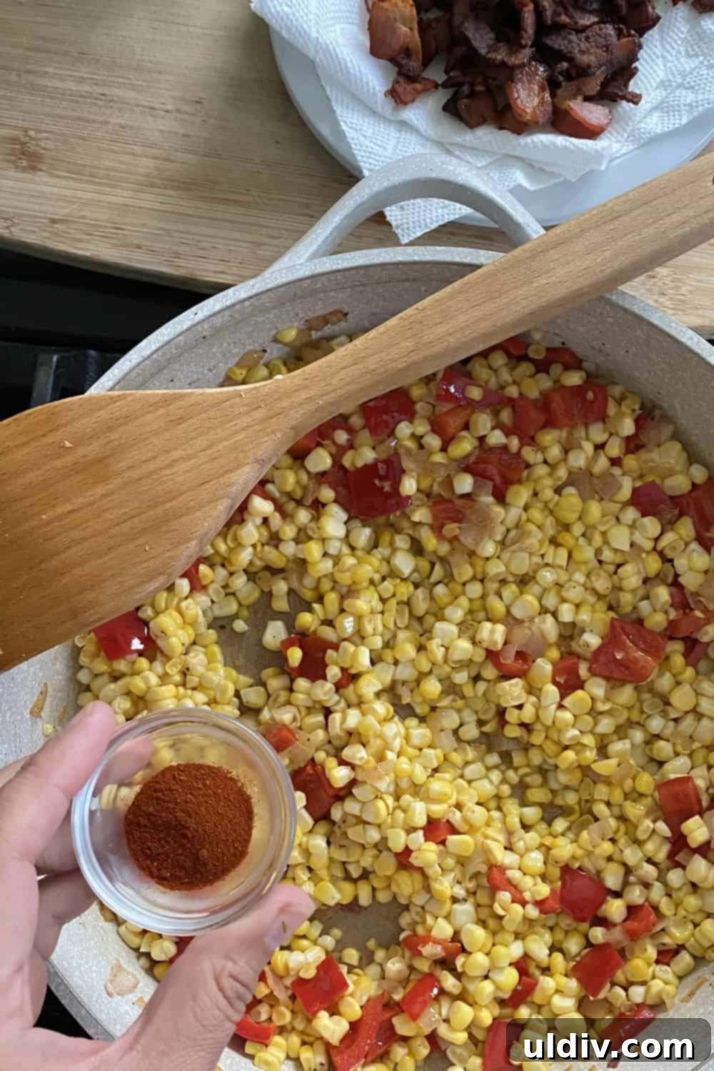 A hand adding smoked paprika to the sautéing sweet corn and vegetable mixture in a pan, infusing it with a rich, smoky flavor.