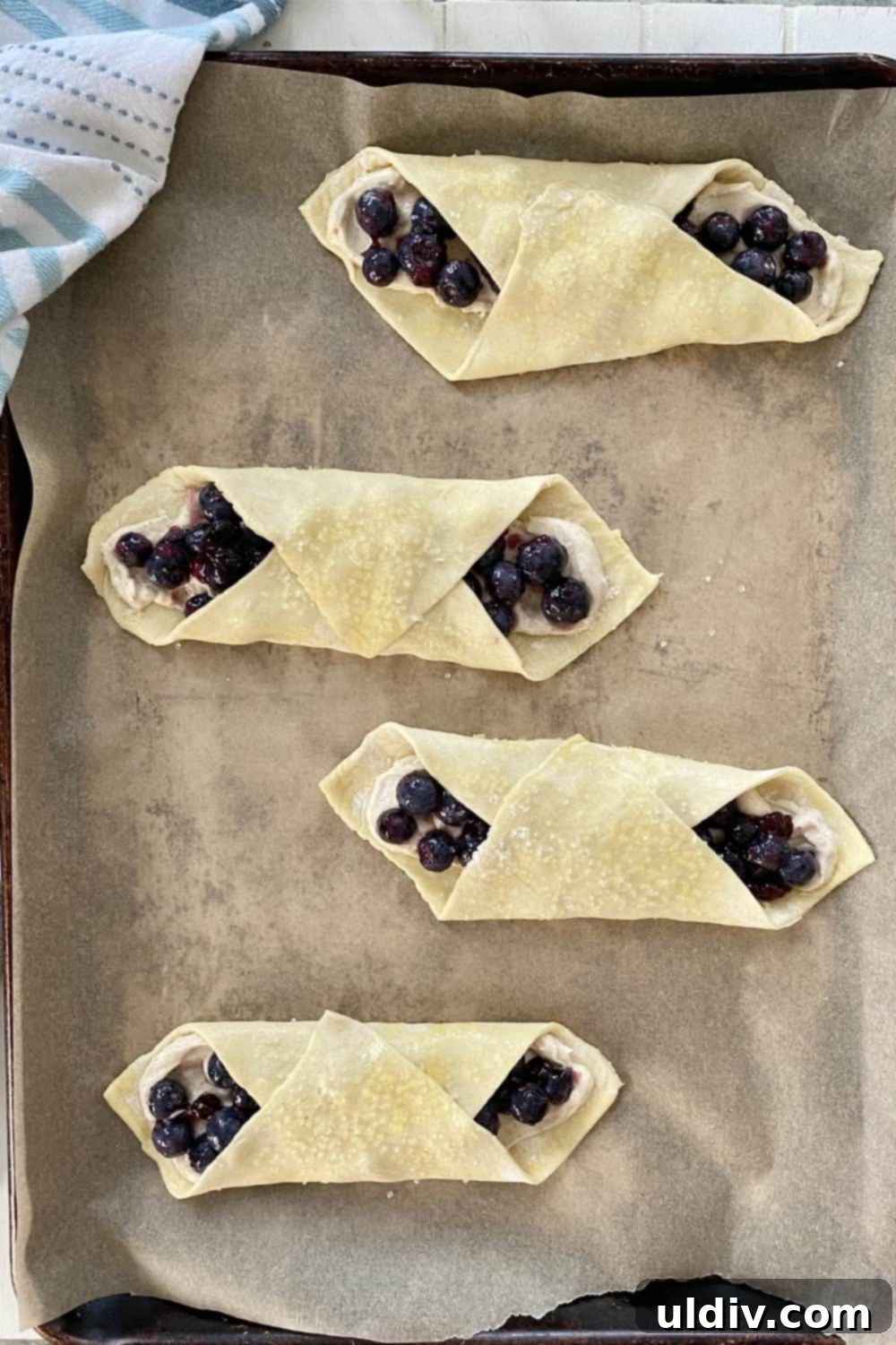 Blueberry Cream Cheese Danish Bliss 8 Unbaked blueberry and cheese danishes, brushed with egg wash and sprinkled with sugar, on a parchment-lined baking sheet.