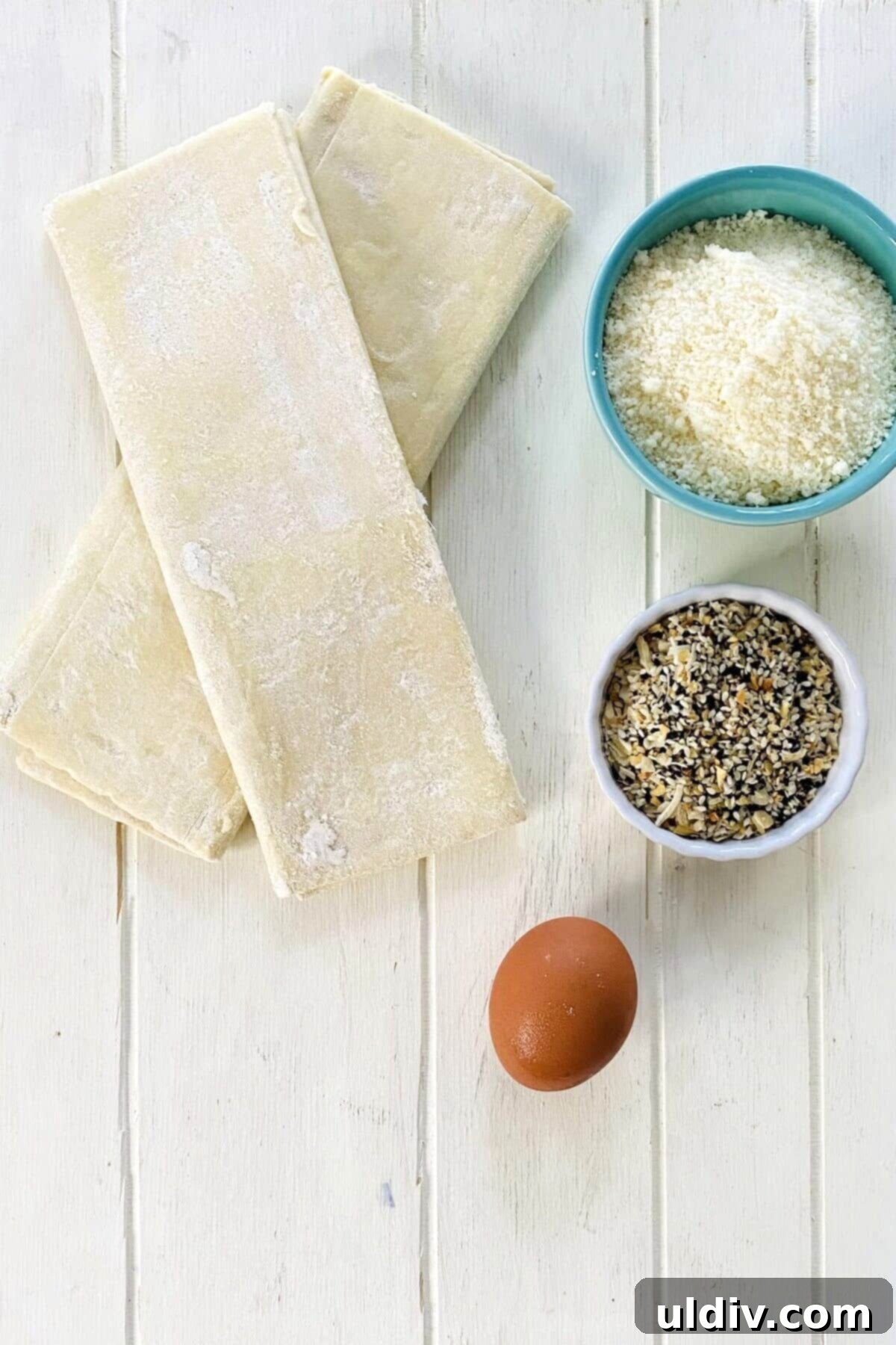 Flaky Everything Bagel Cheese Twists 3 Puff pastry sheets, everything bagel seasoning, grated Parmesan cheese, and an egg laid out as ingredients for cheese twists.
