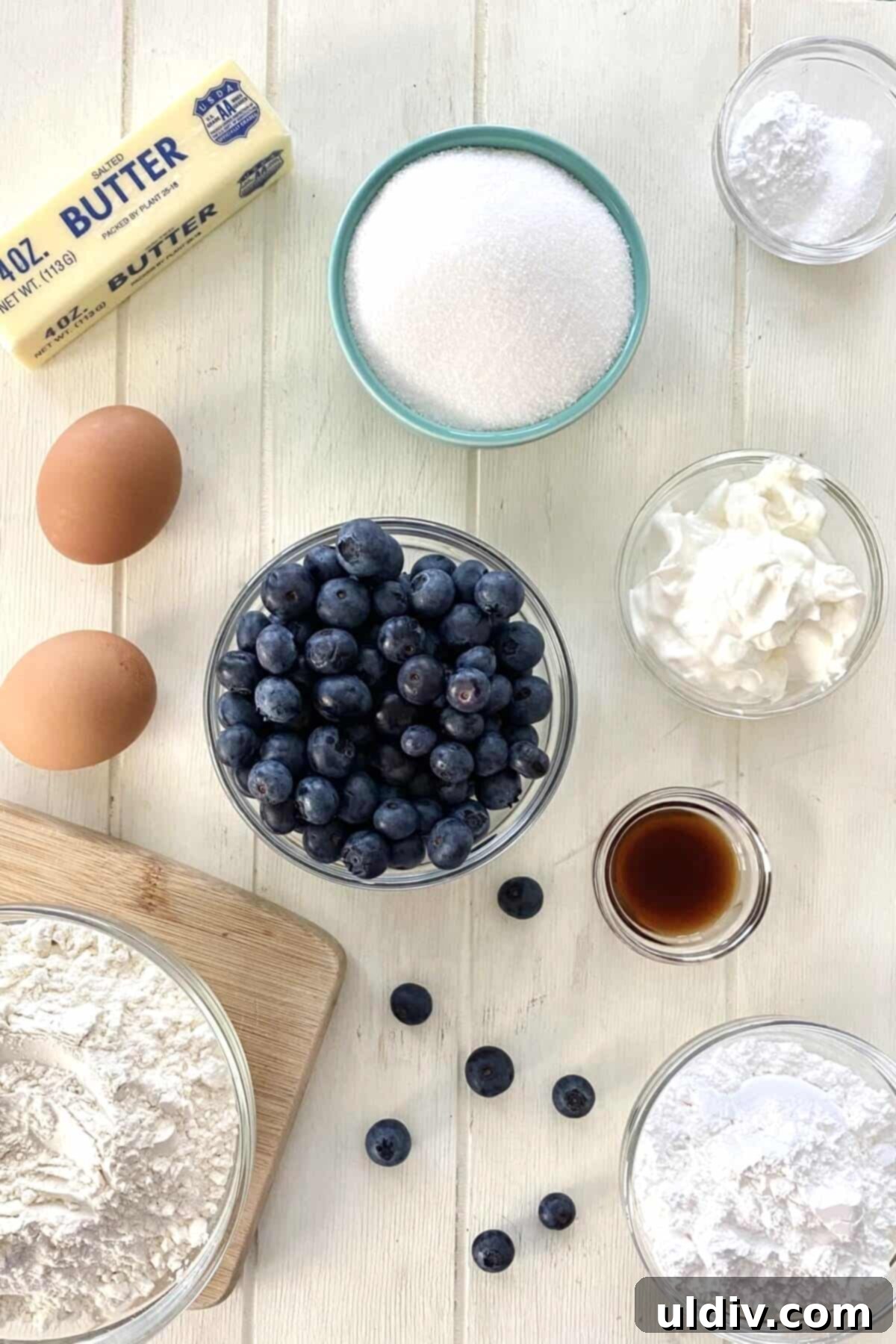 Bursting Blueberry Muffin Cake 3 A visual display of key ingredients: butter, flour, fresh blueberries, sugar, eggs, sour cream, and buttermilk, neatly arranged for a blueberry muffin cake recipe.