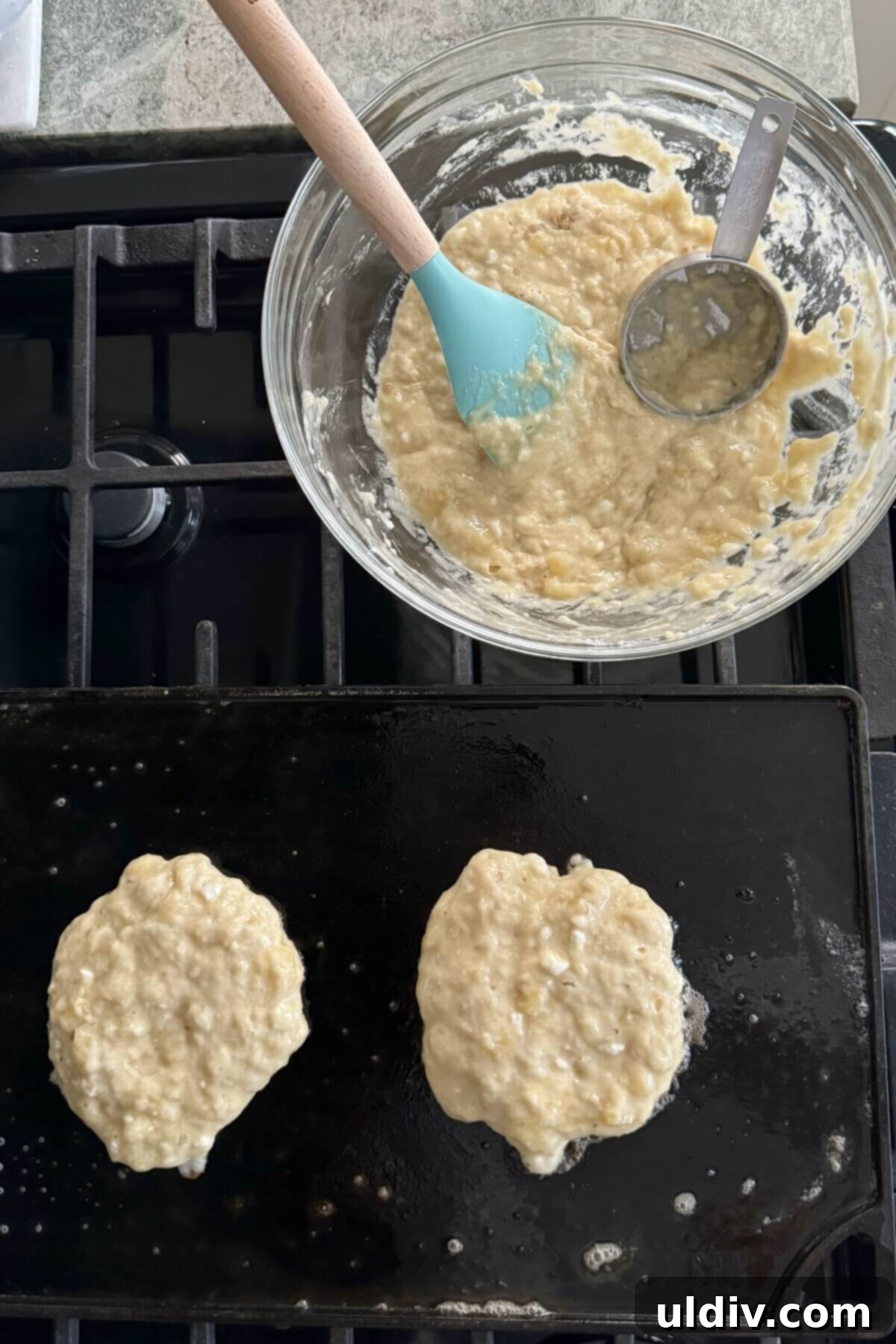 Power-Packed Banana Cottage Cheese Protein Pancakes 10 Two cooking pancakes and a scoop of batter on a hot griddle, showing the start of the cooking process.