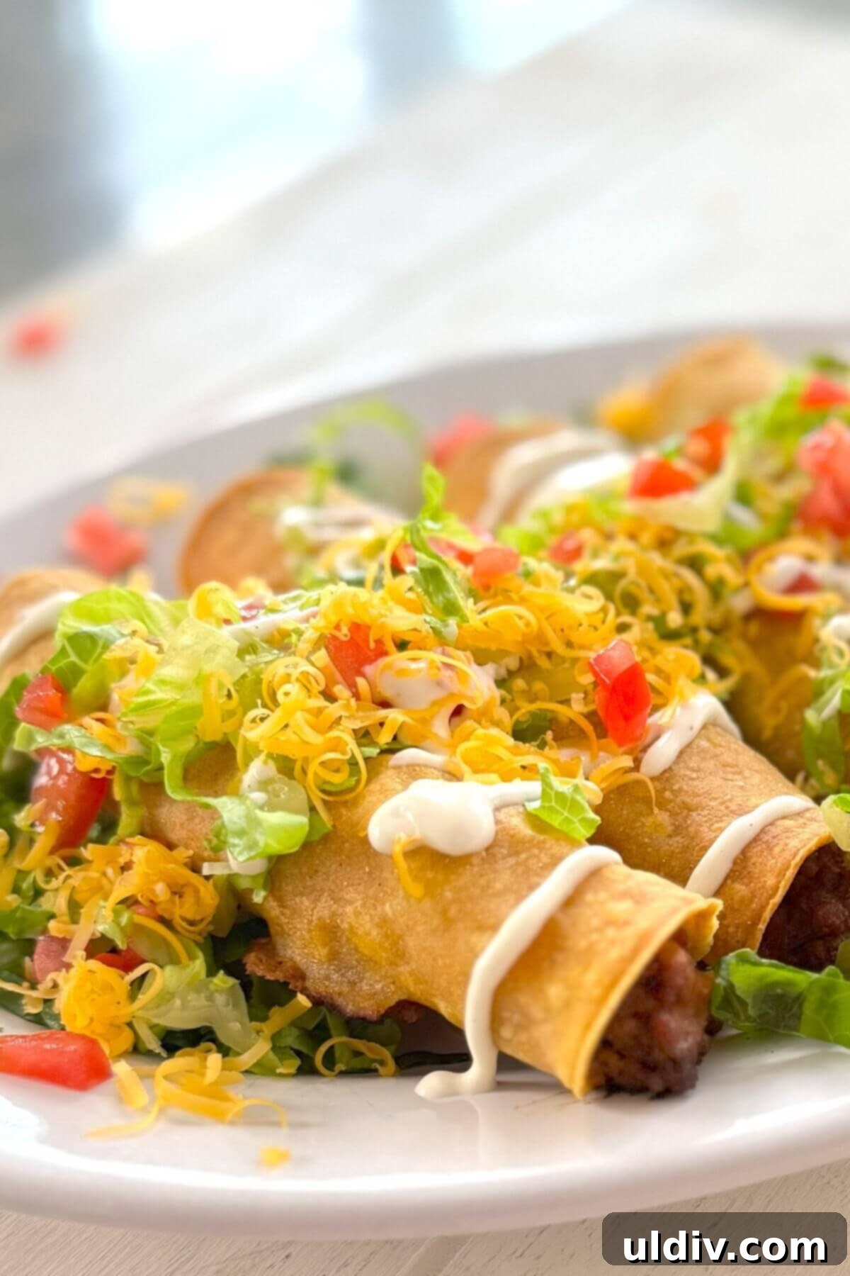 Crunchy Baked Taquitos 14 A vibrant plate of rolled tacos garnished with fresh sour cream, shredded cheese, crisp lettuce, and diced tomatoes, ready to be enjoyed.