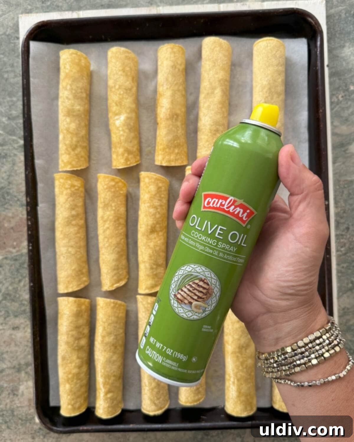 Crispy Baked Taquitos 10 Rolled tacos on a baking sheet being lightly coated with cooking spray for crispiness.