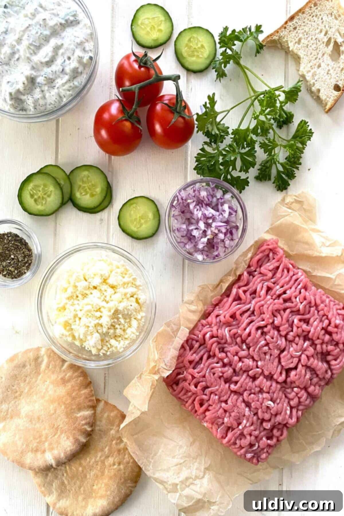 A colorful array of fresh ingredients for Greek turkey burgers: ground turkey, chopped onion, cucumber, tomatoes, feta cheese, and mini pita bread.