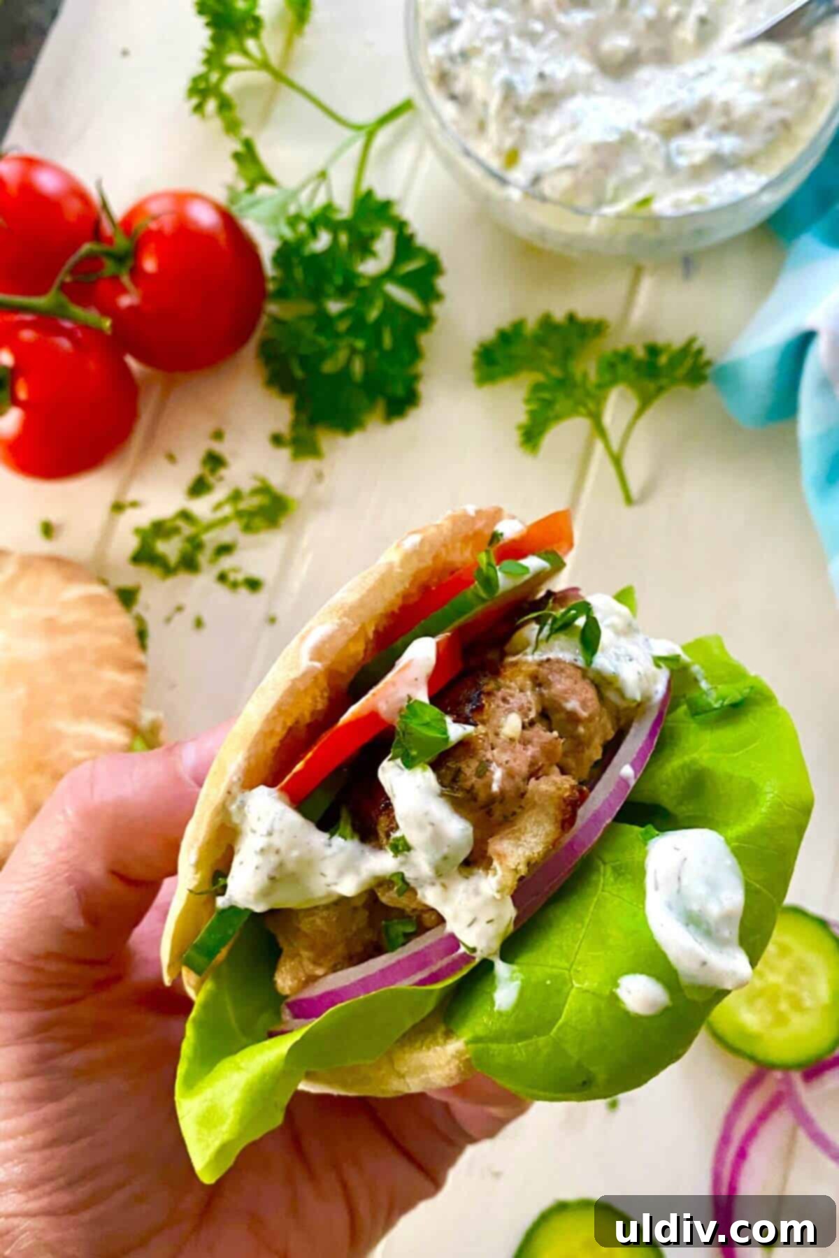 A hand holding a perfectly assembled Greek turkey burger in a mini pita with fresh toppings and tzatziki.