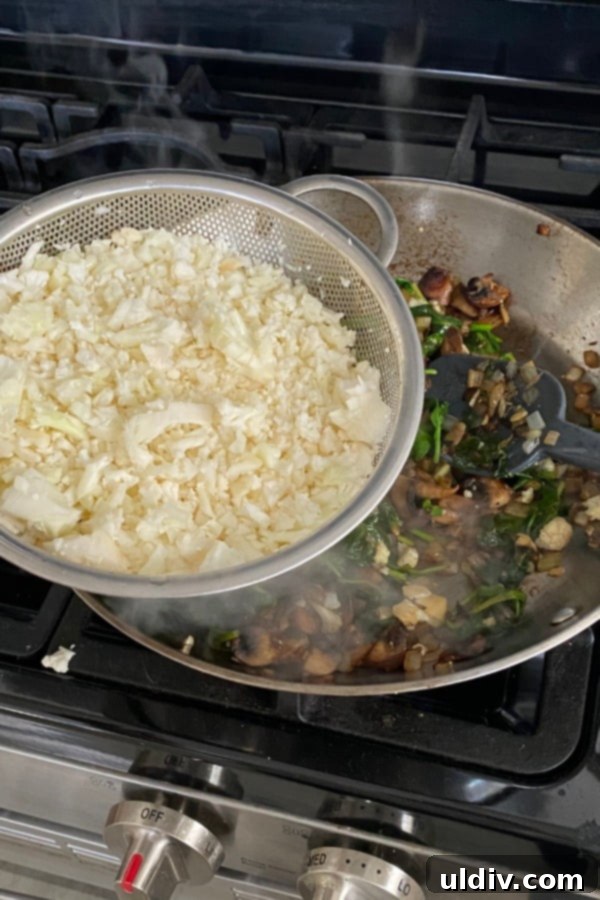 Easy 20-Minute Cauliflower Pilaf 5 A close-up view of a sauté pan, showing riced cauliflower being added to a mixture of already cooked mushrooms, onions, and wilted spinach, ready to be combined.