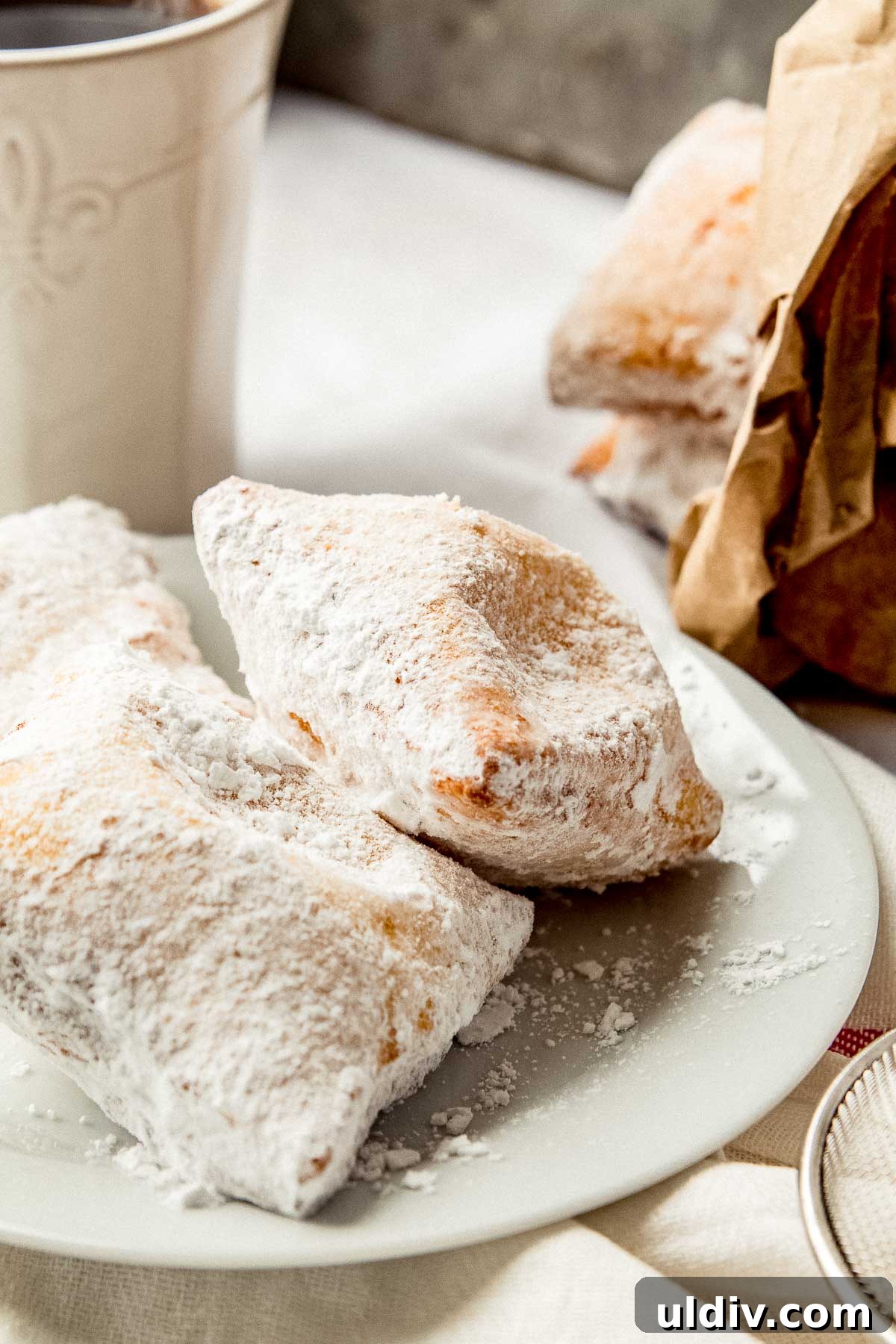 New Orleans Beignets: Classic Café-Style Powdered Sugar Treats 3 Beignets on a plate with a cup of coffee on the side.